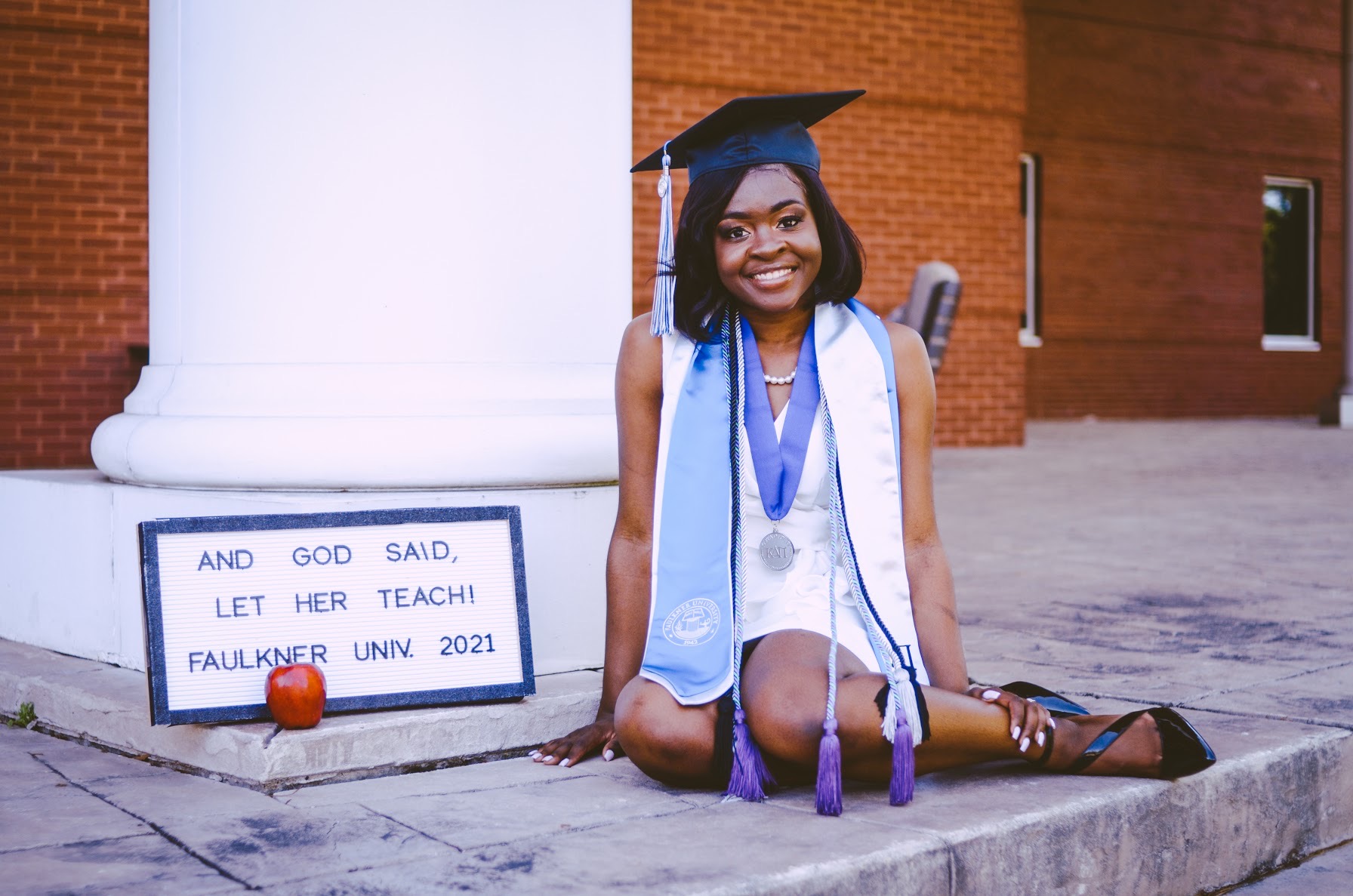 Zanesha Rhodes sits on stone steps, by a white pillar in Faulkner regalia with an apple and a sign that reads, "And God said Let her Teach. Faulkner University 2021."